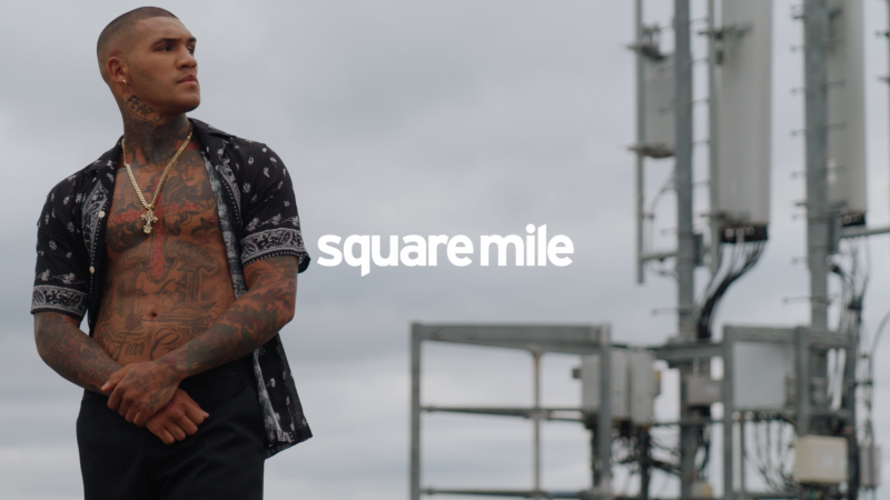 Tattooed man in open shirt with necklace looks left against a grey sky with radio towers. Text reads 'square mile'.