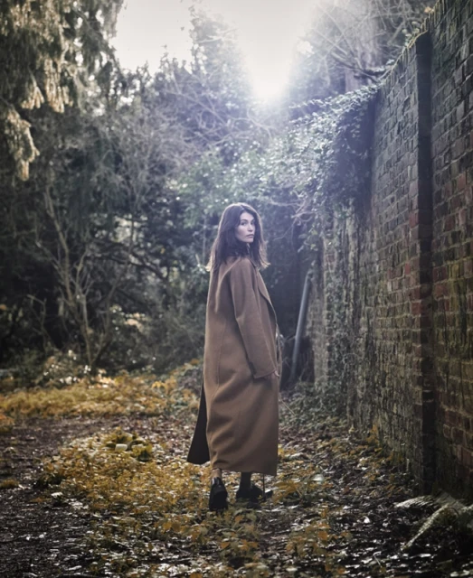 A woman in a brown coat stands on a path next to a weathered brick wall, surrounded by trees in a naturally lit outdoor setting.