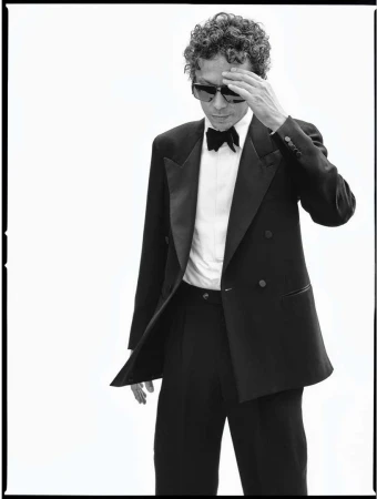 Black and white portrait of an adult male in a tuxedo and sunglasses, standing against a white backdrop, hand to his head.