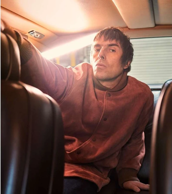 Adult male in a reddish jacket sitting inside a car, looking directly at the camera with a serious expression, interior shot.
