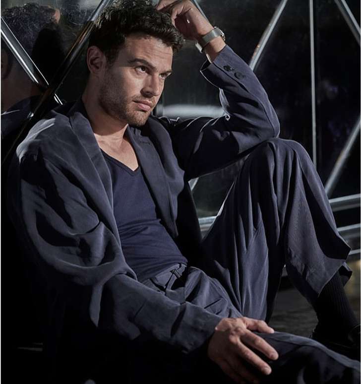 Portrait of a handsome man sitting on the floor in a suit, striking a thoughtful pose in front of a reflective background.