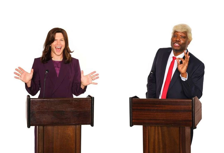 Two people dressed as politicians stand behind podiums on a white background. One person is dressed as a woman, the other as Trump.