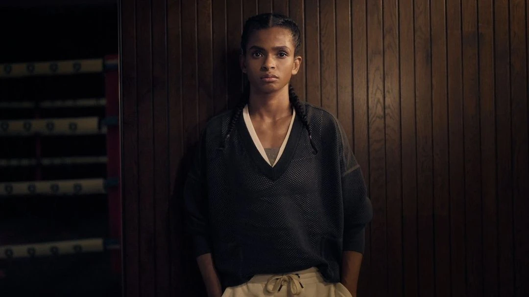 A young woman with braided hair stands in a locker room with a determined expression, wearing a dark athletic shirt and light sweatpants.