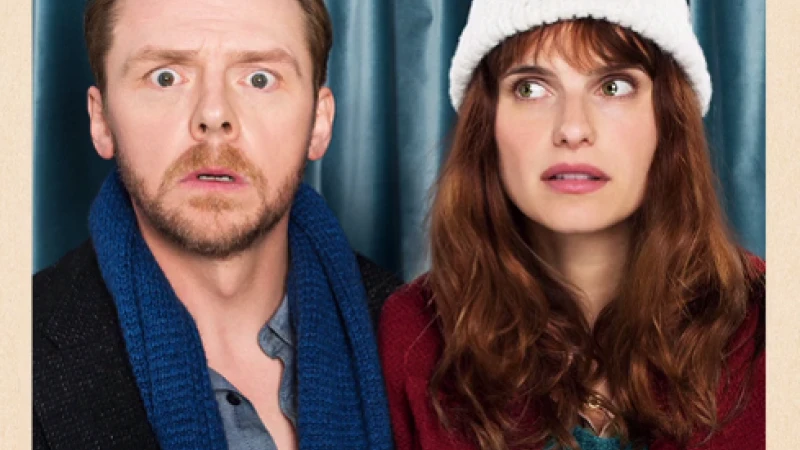 Eye-level shot of Simon Pegg with a surprised expression and Lake Bell wearing a white hat, posing against a blue curtain backdrop.