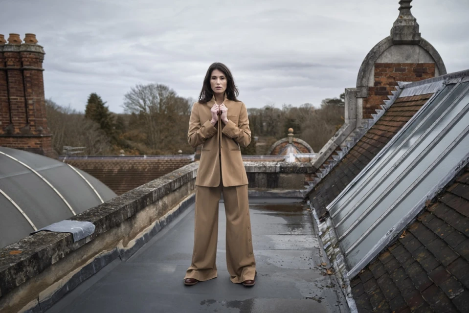 Woman in tan suit stands on rooftop with a cloudy sky. She is the primary subject, with building features and trees in background.