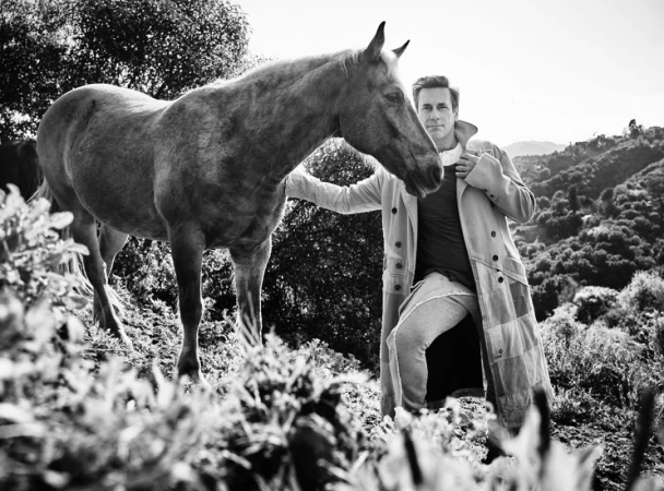 Man in long coat standing outdoors next to a horse in a grassy hilly landscape captured in monochrome