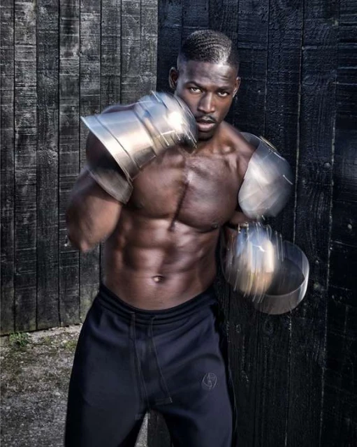 Muscular Black man in athletic wear and metallic arm guards punches toward the camera against a dark, weathered wood wall.