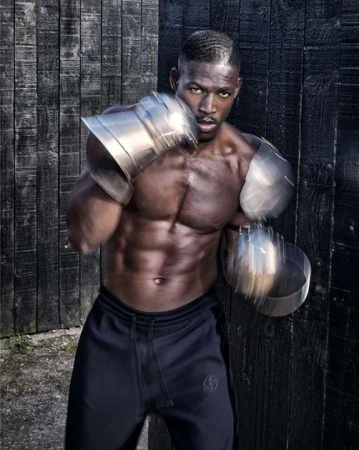 Muscular Black man in athletic wear and metallic arm guards punches toward the camera against a dark, weathered wood wall.