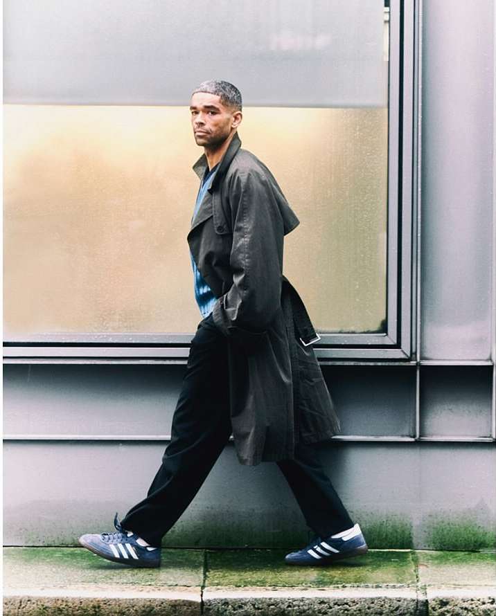A man in a trench coat walking past a modern glass and metal building. He wears dark pants and Adidas Sambas in an urban environment.