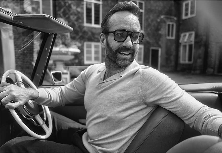 Black and white portrait of a man driving a vintage convertible car, smiling while wearing glasses and a light-colored long sleeve shirt.