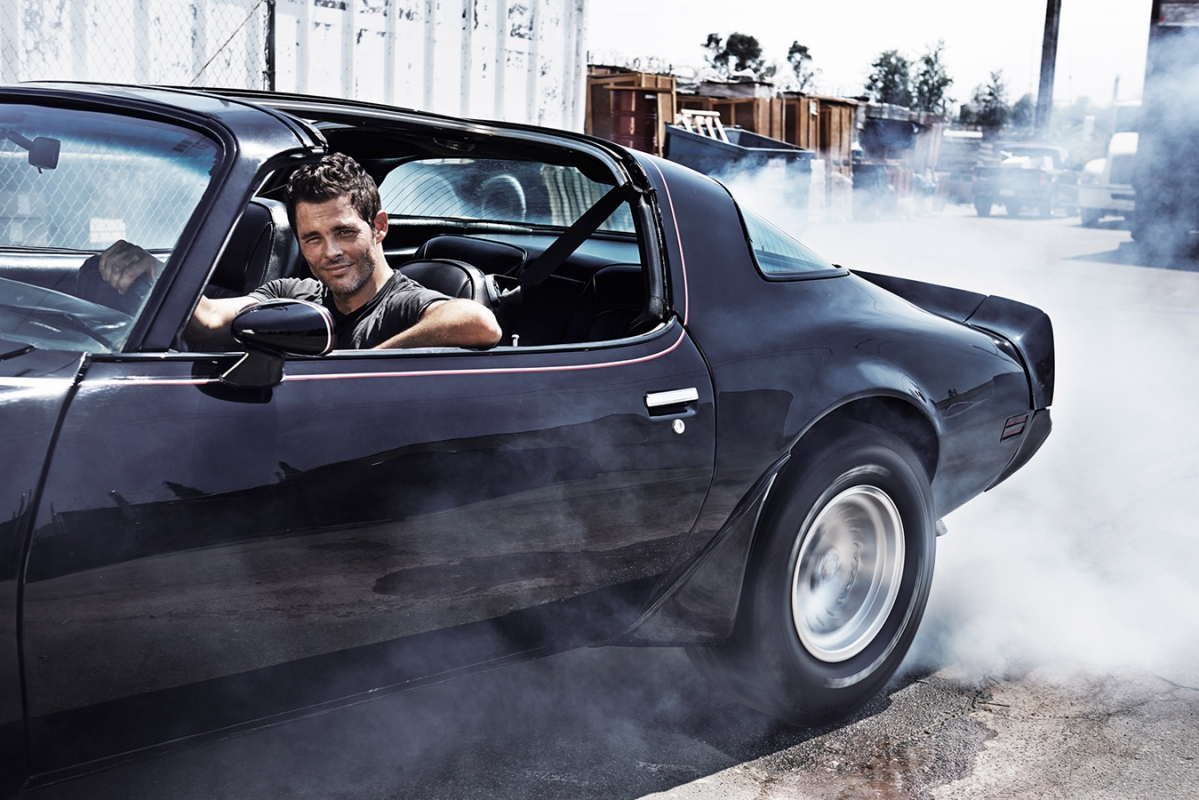 Man in a classic black sports car with smoke billowing from the tires in an outdoor industrial setting, capturing a dynamic automotive scene.