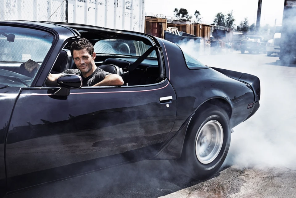 Man in a classic black sports car with smoke billowing from the tires in an outdoor industrial setting, capturing a dynamic automotive scene.