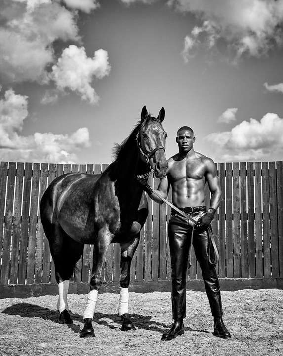 Striking black and white shot of a muscular man in leather pants standing confidently with a horse in an outdoor enclosure.