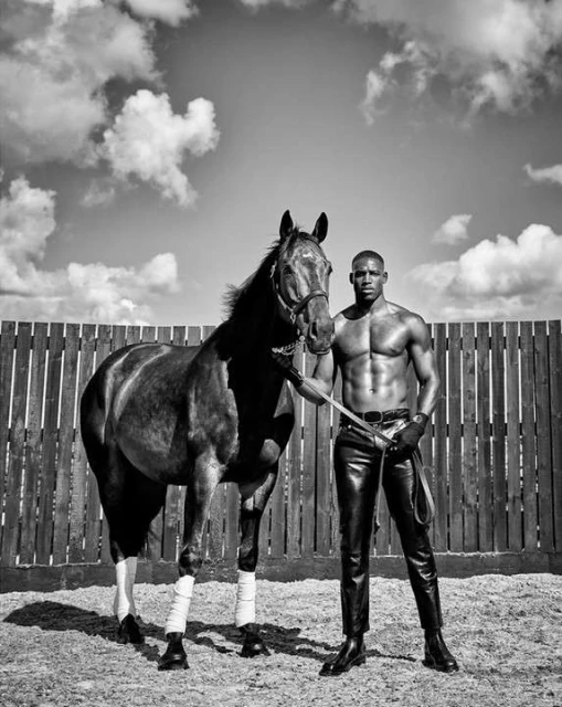 Striking black and white shot of a muscular man in leather pants standing confidently with a horse in an outdoor enclosure.