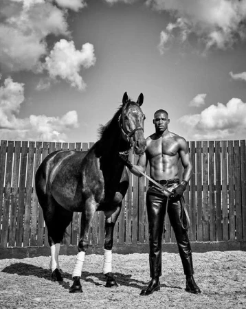 Striking black and white shot of a muscular man in leather pants standing confidently with a horse in an outdoor enclosure.