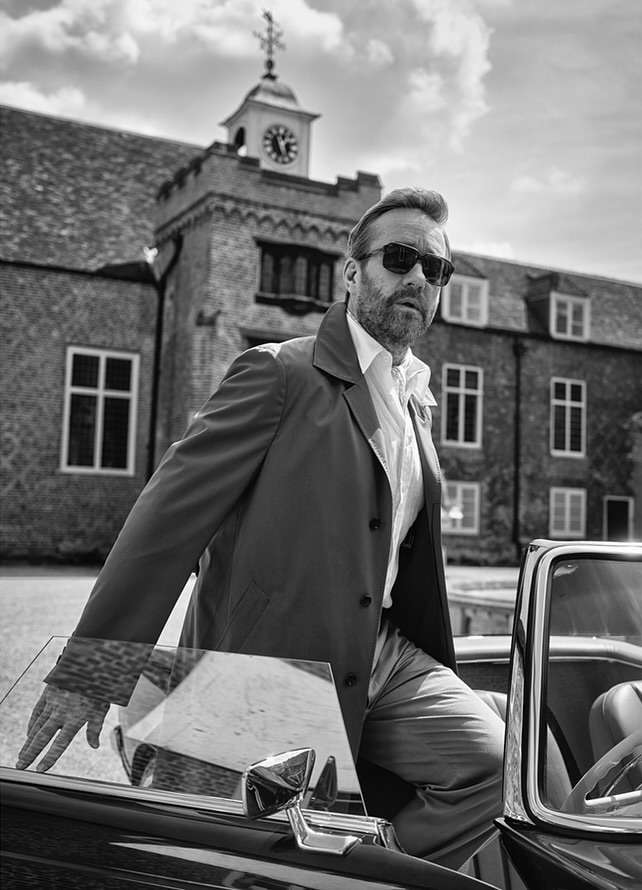 Mature man with sunglasses exits vintage convertible in front of brick building with clock tower. Black and white fashion portrait.