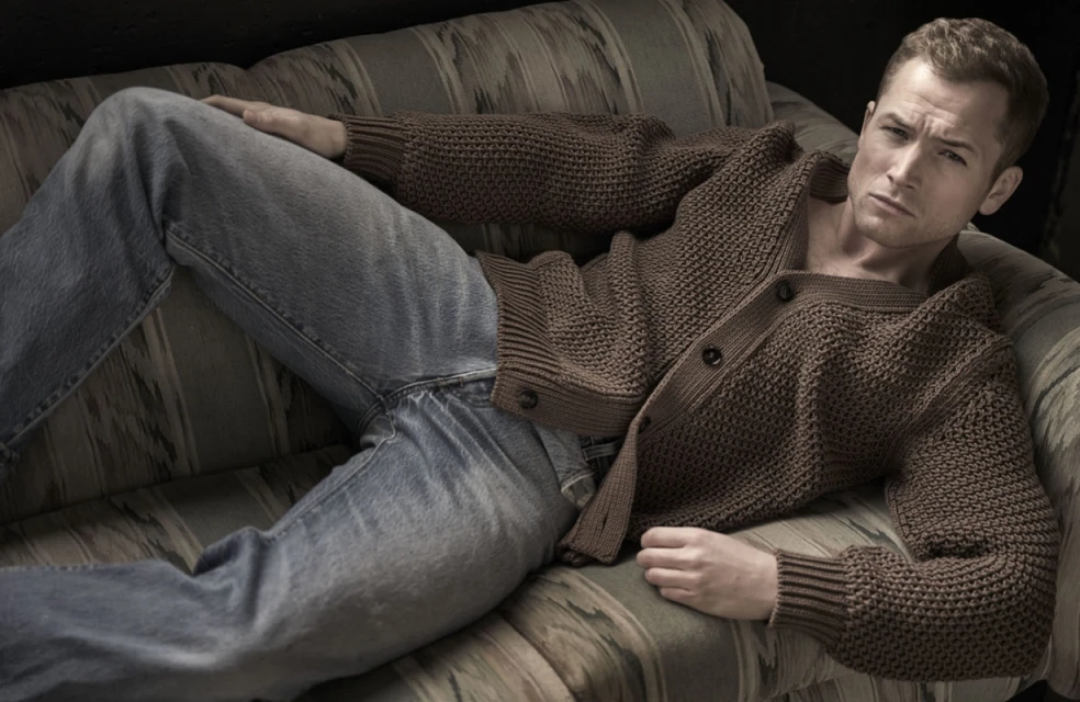 Adult male reclining on a couch in a brown knitted cardigan and blue jeans in an indoor setting with dark background.