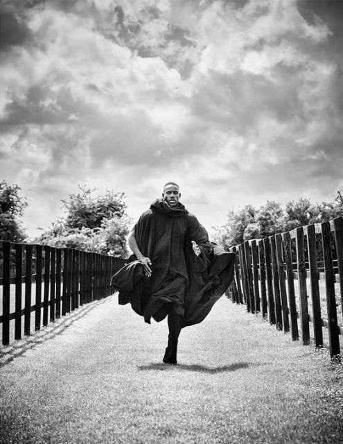 Black and white portrait of a Black male model running on a path through a park, wearing a dark cloak under a cloudy sky.