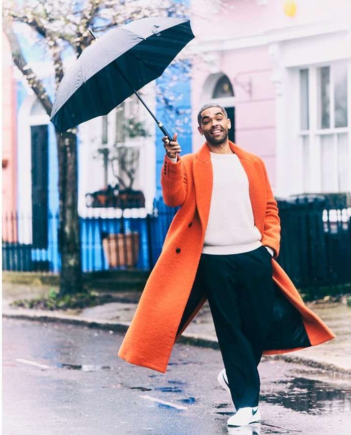 A man in an orange coat holds a black umbrella outdoors on a wet day, standing in front of colorful houses on a city street.