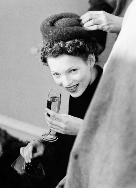 Monochrome image of a smiling woman with curly hair and elaborate headwear, holding a champagne glass, with a partial view of a person nearby.