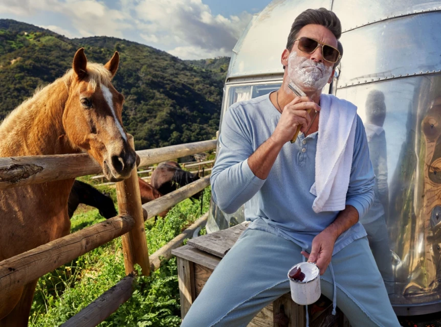 Mature man with shaving cream and straight razor sits next to a horse in a rural outdoor setting with mountains in the background.