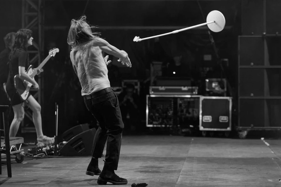 Black and white concert shot of a shirtless male singer with long hair performing on stage with a guitarist and audio equipment.