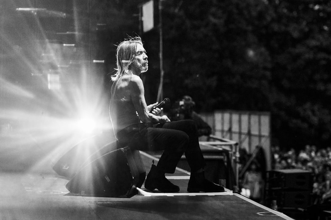 Monochrome concert image of a shirtless male singer sitting on a speaker with a microphone, captured in a dramatic, high-contrast black and white style.
