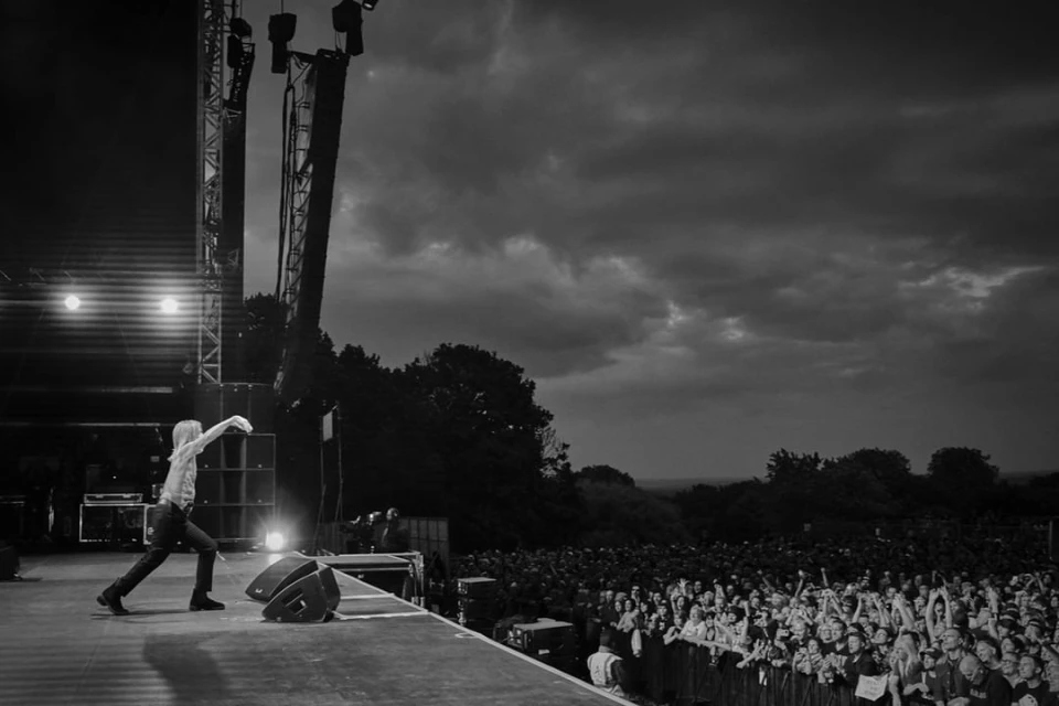 Black and white shot of a male singer performing on a stage in front of a large crowd with stage lighting and a cloudy sky overhead.