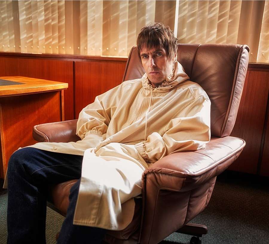 Adult male sitting in a leather chair wearing a beige hooded jacket with an unsmiling expression in an indoor wood-paneled setting.