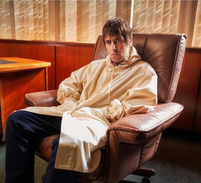 Adult male sitting in a leather chair wearing a beige hooded jacket with an unsmiling expression in an indoor wood-paneled setting.