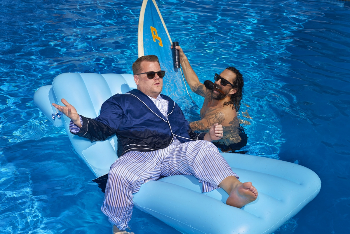 Two men in a pool, one lounging on a blue inflatable, the other holds up a spray bottle against a bright blue water background.