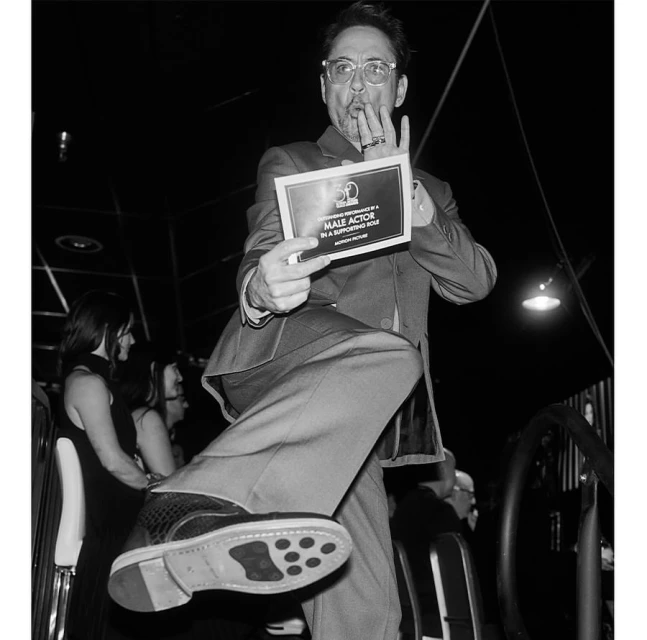 Black and white photo of an adult male actor holding an award plaque, wearing a suit, and playfully stepping forward with his leg up.