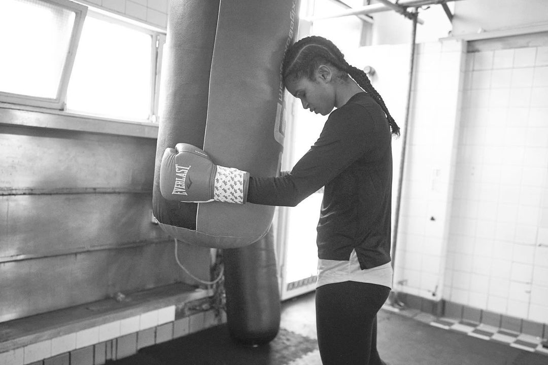 A boxer rests her head against a heavy bag in a gym with Everlast gloves and hand wraps. The image is black and white, reflecting weariness.