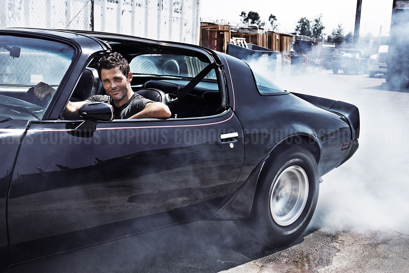 Adult male driving a black vintage sports car with smoke billowing around the wheels in an outdoor industrial environment.