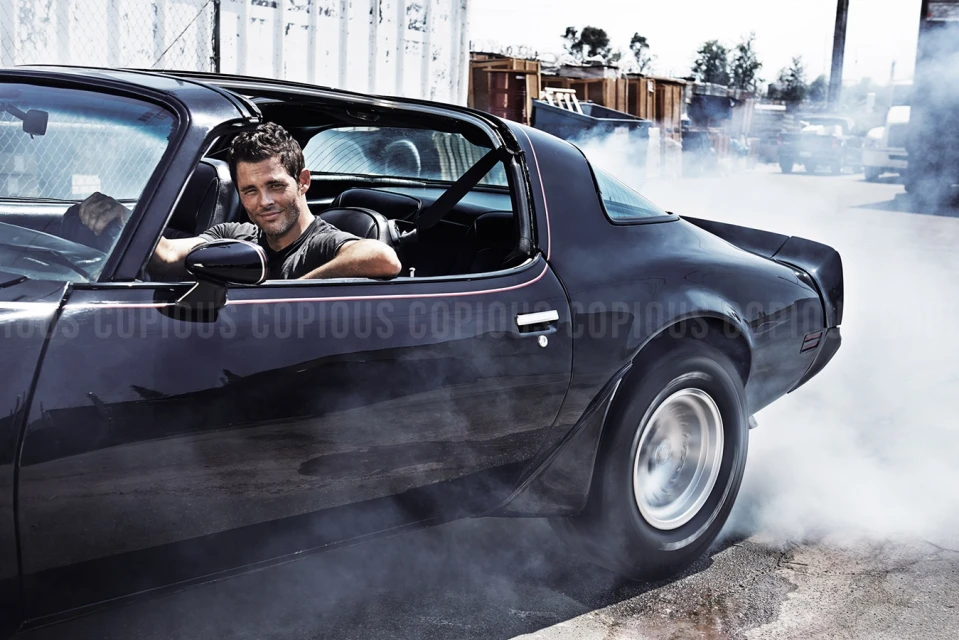 Adult male driving a black vintage sports car with smoke billowing around the wheels in an outdoor industrial environment.