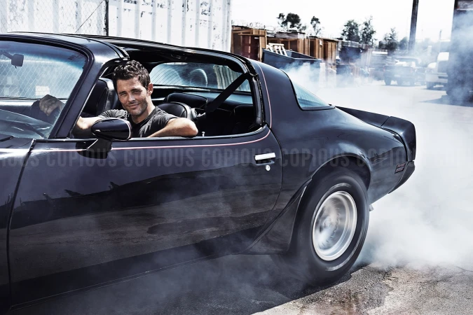 Adult male driving a black vintage sports car with smoke billowing around the wheels in an outdoor industrial environment.