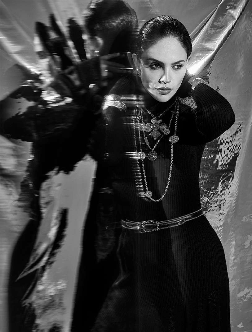 Black and white fashion portrait of a woman wearing a ribbed turtleneck dress, silver belt, and multiple silver necklaces against foil backdrop.