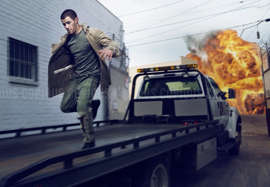 Adult male runs from a large explosion on a truck flatbed in an urban alleyway with buildings and telephone wires overhead.