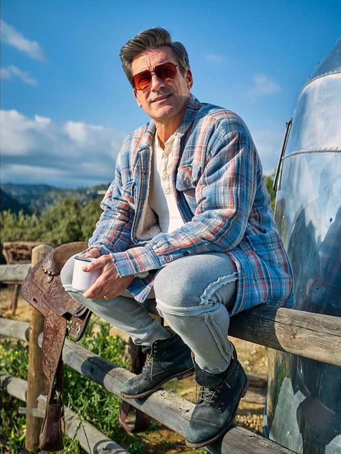 Man sitting on a wooden fence next to a silver trailer, holding a white mug, wearing jeans, boots, and sunglasses in a rural outdoor setting.