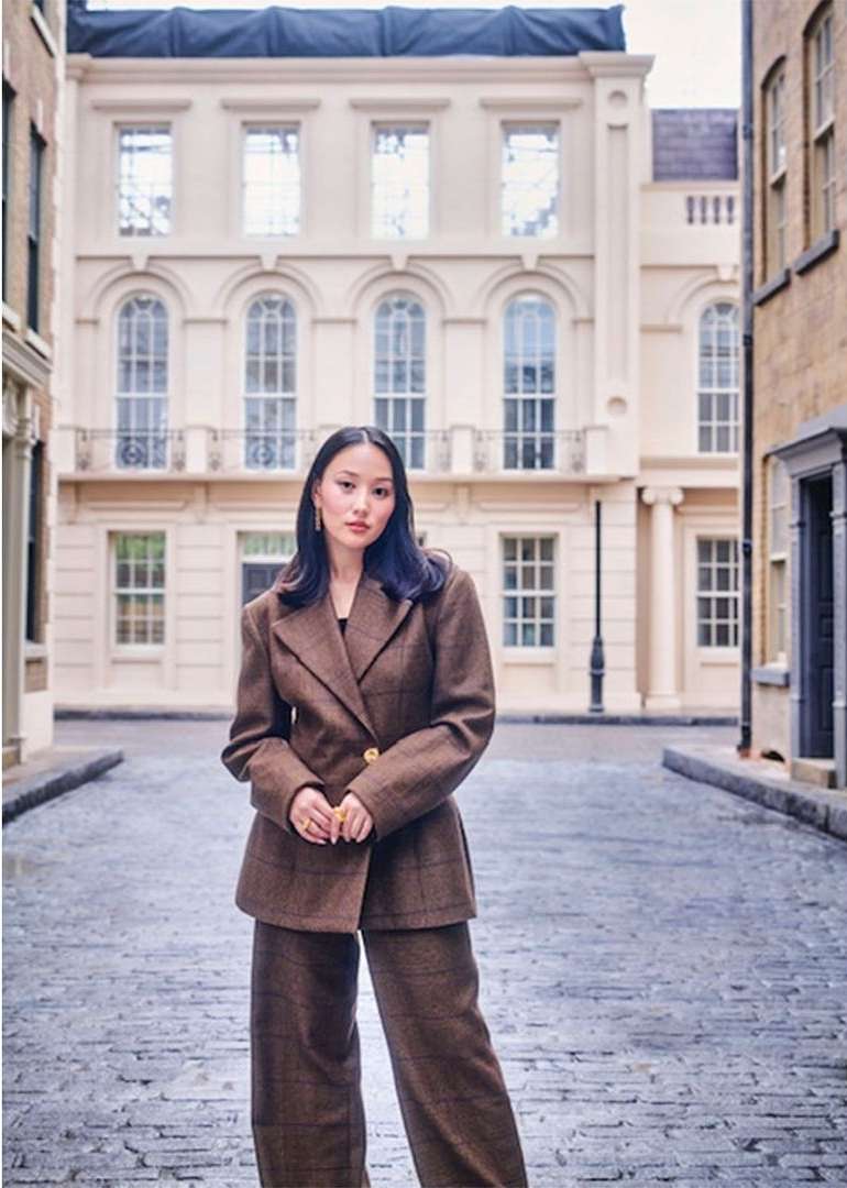 Woman in a brown pantsuit stands on a cobblestone street in front of a cream-colored building with arched windows and a black lamppost.