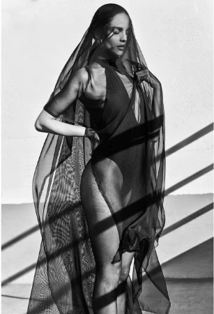 Monochrome portrait of a woman in a sheer black veil and bodysuit standing against a textured wall with strong diagonal shadows.