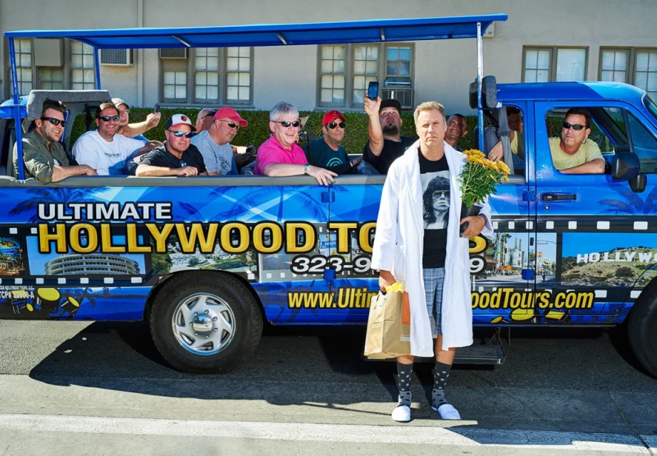 Man in a lab coat stands outside a blue Hollywood tour van filled with tourists on a sunny day, holding flowers and a bag.