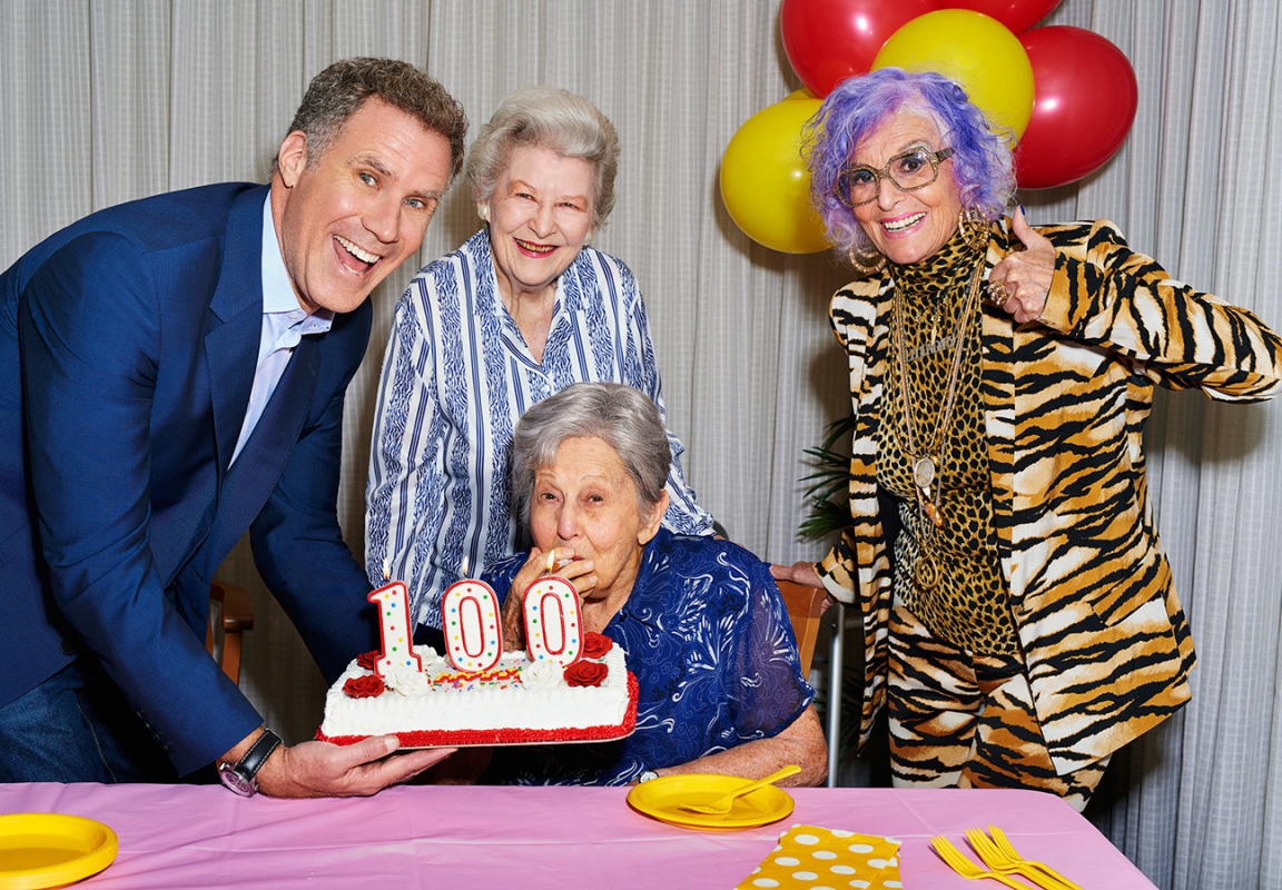Group of people celebrating a 100th birthday with a cake with candles, balloons, and a table covered in a pink tablecloth.