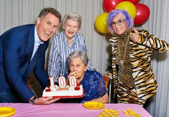 Group of people celebrating a 100th birthday with a cake with candles, balloons, and a table covered in a pink tablecloth.