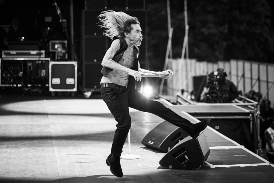 Live concert photo of a male performer kicking one leg up on stage with speakers, performing in front of a dark background and stage lights.