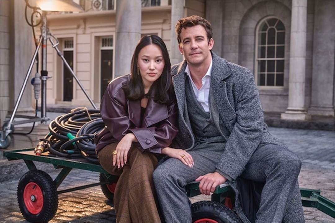 Outdoor portrait of a smiling man and woman in stylish coats sitting on film equipment. Grey stone building and columns in the background.