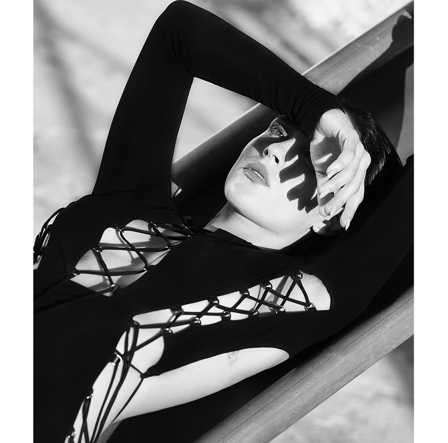 Black-and-white portrait of a woman with short hair, wearing a long-sleeved lace-up top, posing outdoors in dappled sunlight.