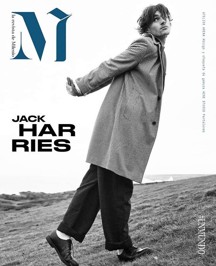 Black and white fashion editorial shot of a male model in a patterned coat and dark pants posing on a grassy hill for a magazine cover.