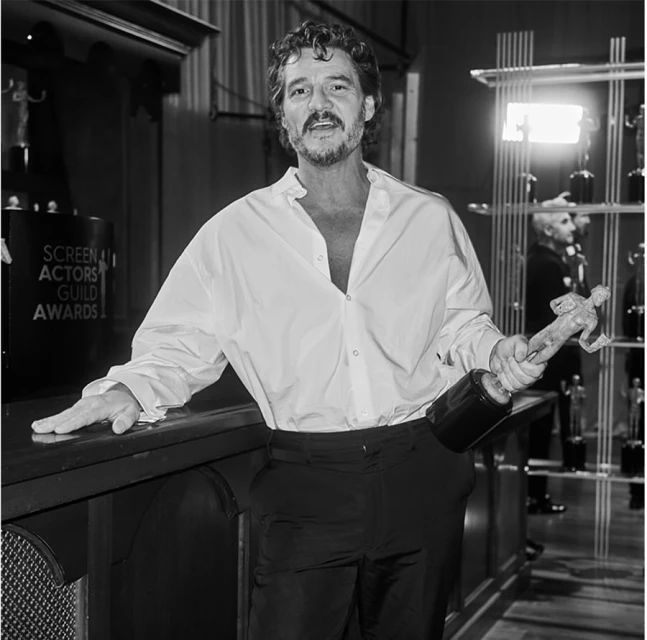 Black and white photo of Pedro Pascal in an unbuttoned white shirt, holding a Screen Actors Guild award, with SAG awards signage in the background.