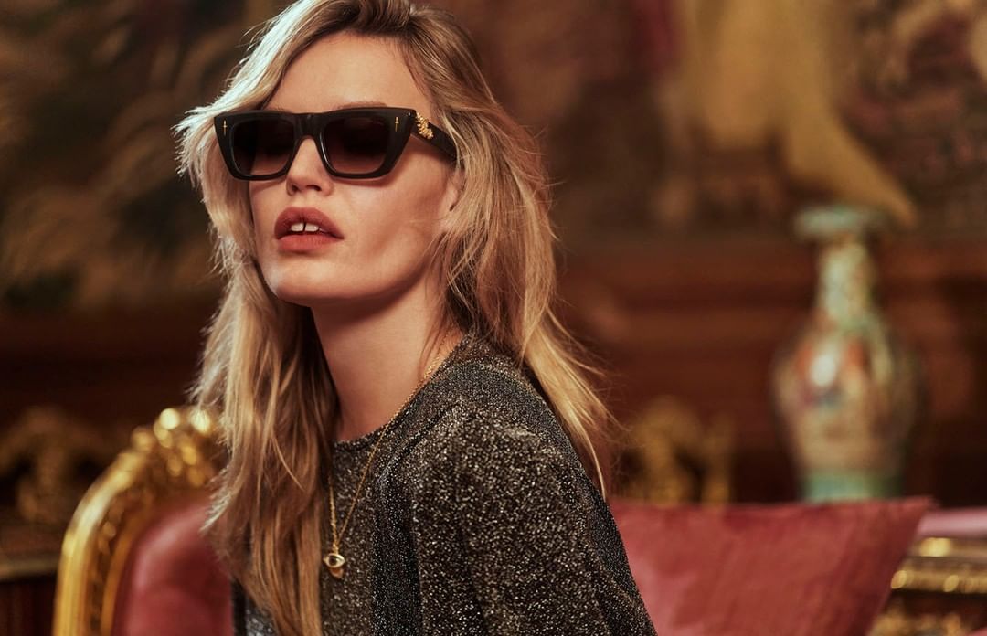 Adult female wearing designer sunglasses and a shimmering black blouse, gazing at the camera in a luxurious indoor setting.